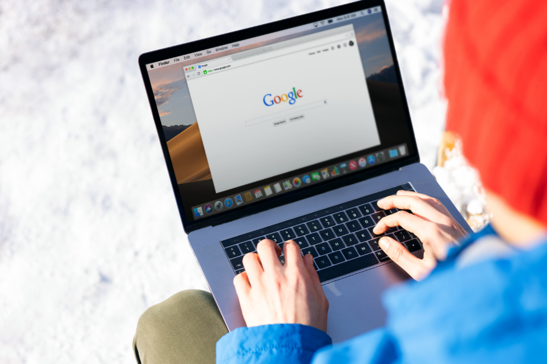 A person using Google Keyword Planner on a laptop, analyzing search volume and competition metrics. The screen displays keyword suggestions with popularity bars. A notebook and coffee cup are on the desk.